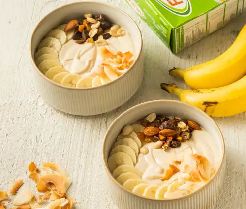 Two white ceramic bowls filled with yogurt, sliced bananas, almonds, raisins, and toasted coconut flakes.