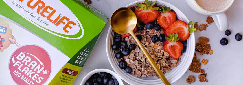 A white bowl filled with bran flakes, blueberries, and sliced strawberries, topped with a golden spoon.
